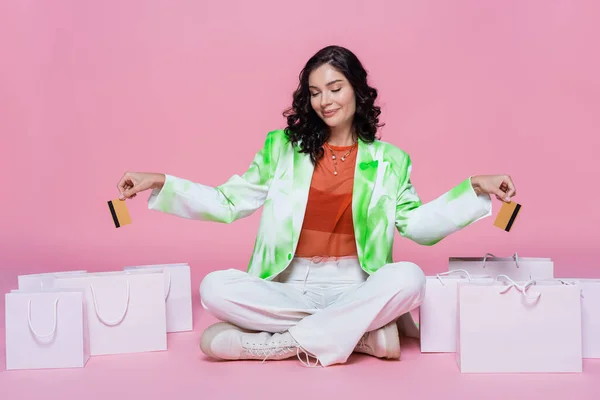 Pleased woman in green blazer holding credit cards while sitting with crossed legs near shopping bags on pink — Stock Photo