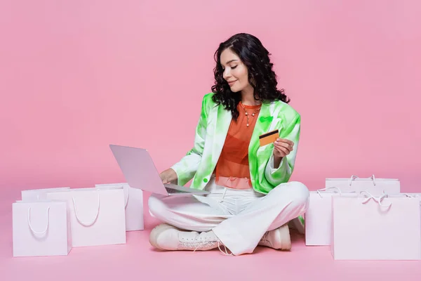 Mujer complacida en blazer usando el ordenador portátil mientras sostiene la tarjeta de crédito cerca de bolsas de compras en rosa - foto de stock