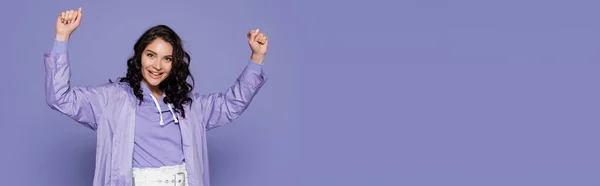 Excited young woman in raincoat standing with raised hands isolated on purple, banner — Stock Photo