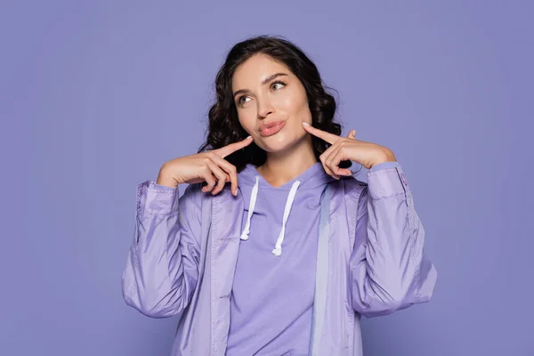 Brunette young woman in raincoat pouting lips and pointing at cheeks isolated on purple — Stock Photo