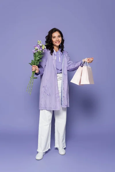 Pleine longueur de jeune femme heureuse en imperméable tenant bouquet de fleurs et sacs à provisions sur violet — Photo de stock