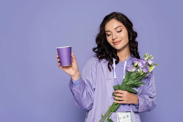 Happy young woman in raincoat holding bouquet of flowers and paper cup isolated on purple — Stock Photo