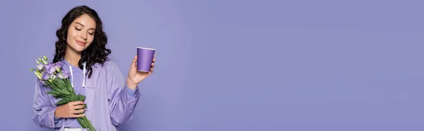 Heureuse jeune femme en imperméable tenant bouquet de fleurs et tasse en papier isolé sur violet, bannière — Photo de stock