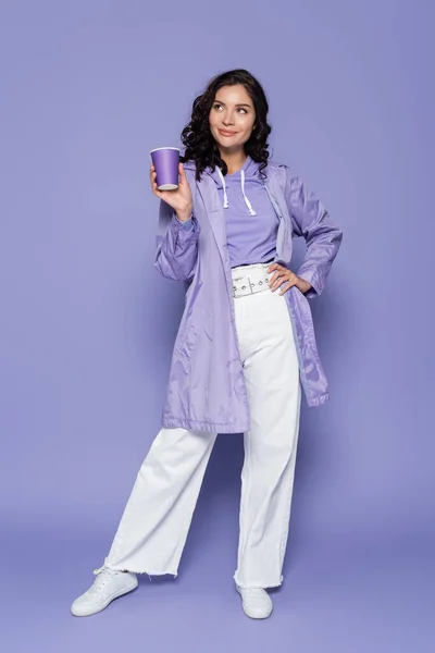 Щаслива молода жінка в плащі тримає паперову чашку, стоячи з рукою на стегні на фіолетовому — стокове фото