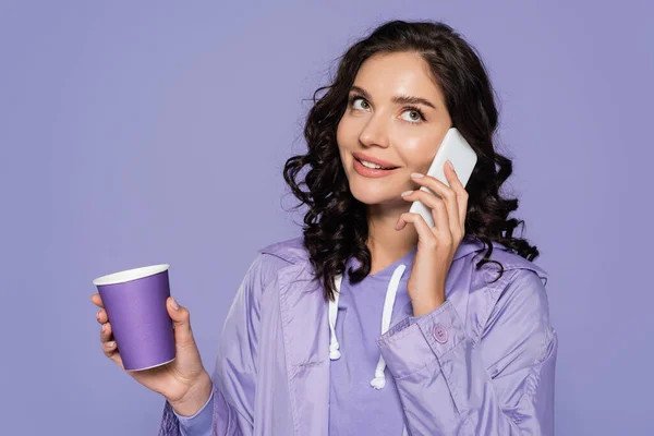 Heureuse jeune femme en imperméable tenant tasse en papier et parlant sur smartphone isolé sur violet — Photo de stock