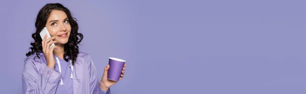 Felice giovane donna in impermeabile in possesso di tazza di carta e parlando su smartphone isolato su viola, banner — Foto stock