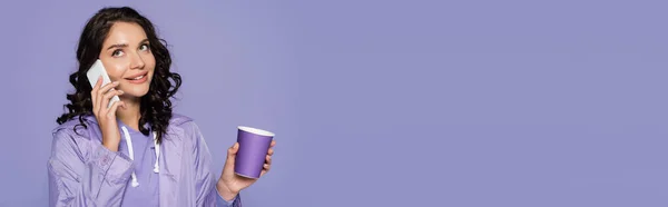 Mujer joven feliz en impermeable sosteniendo taza de papel y hablando en el teléfono inteligente aislado en púrpura, bandera — Stock Photo