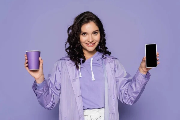 Heureuse jeune femme en imperméable tenant tasse en papier et smartphone avec écran blanc isolé sur violet — Photo de stock