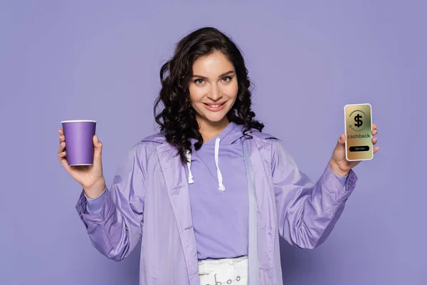 Heureuse jeune femme en imperméable tenant tasse en papier et smartphone avec cashback à l'écran isolé sur violet — Photo de stock