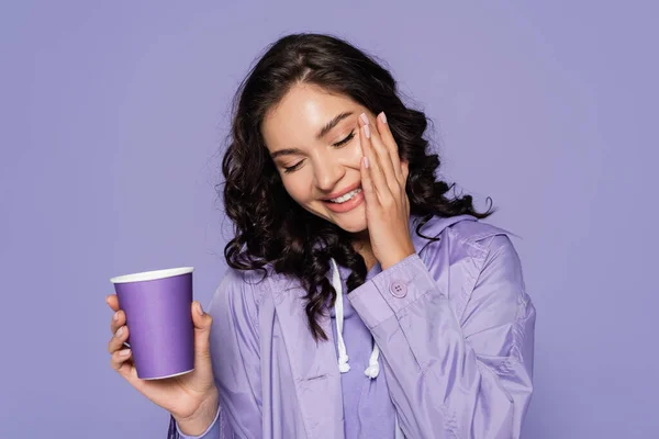 Gioiosa giovane donna in impermeabile contenente tazza di carta isolata su viola — Foto stock