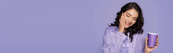 Alegre joven en impermeable sosteniendo taza de papel aislado en púrpura, bandera - foto de stock