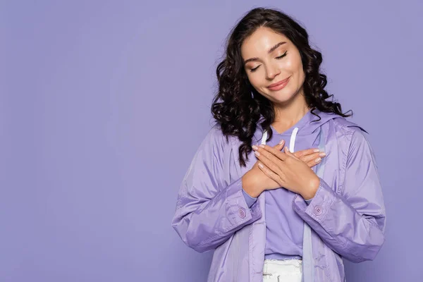 Pleased young woman in raincoat holding hands near chest isolated on purple — Stock Photo