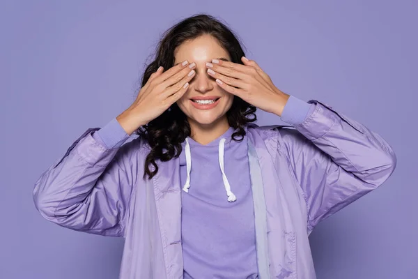 Sonriente joven en impermeable cubriendo ojos aislados en púrpura - foto de stock