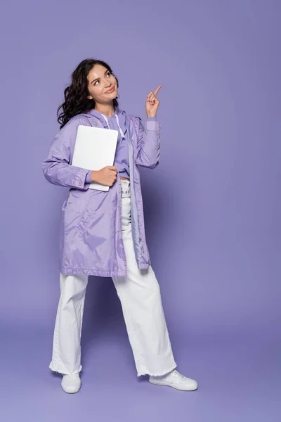 Cheerful young woman in violet raincoat holding laptop and pointing with finger on purple — Stock Photo