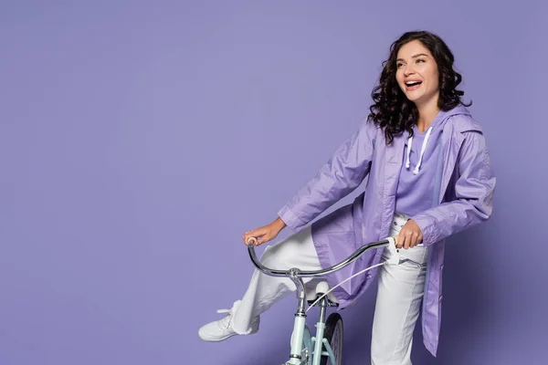Fröhliche junge Frau in violettem Regenmantel sitzt auf einem Fahrrad vereinzelt auf lila — Stockfoto