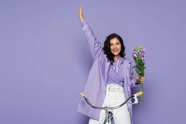 Fröhliche junge Frau in violettem Regenmantel, die Fahrrad fährt und Blumen auf lila hält — Stockfoto