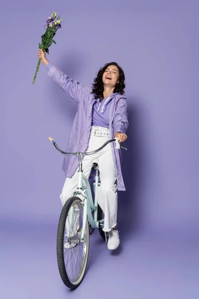 Glückliche junge Frau im Regenmantel, die Fahrrad fährt und Blumen auf lila hält — Stockfoto