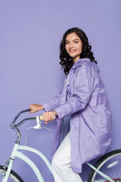 Весела молода жінка в фіолетовому плащі їзда на велосипеді ізольована на фіолетовому — стокове фото