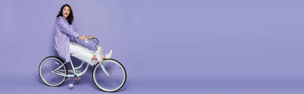 Full length of amazed young woman in violet raincoat riding bicycle on purple, banner — Stock Photo