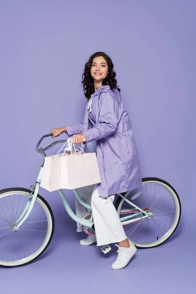 Fröhliche junge Frau in violettem Regenmantel, die Fahrrad fährt und Einkaufstüten auf lila hält — Stockfoto