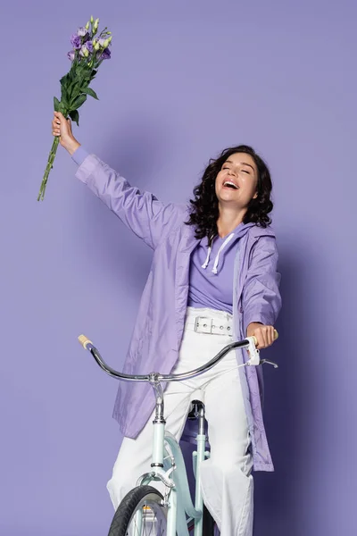 Happy young woman in raincoat riding bicycle and holding eustoma flowers on purple — Stock Photo