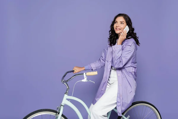 Happy young woman in violet raincoat riding bicycle and talking on smartphone isolated on purple — Stock Photo