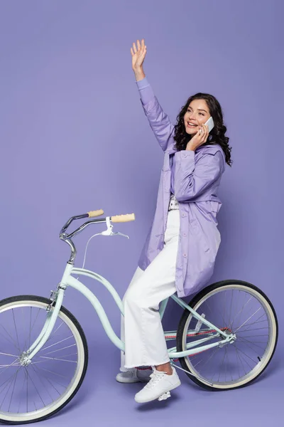 Happy woman in violet raincoat riding bicycle and waving hand while talking on smartphone on purple — Stock Photo