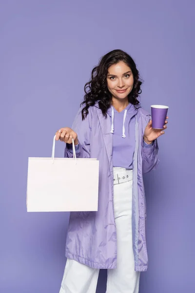 Mujer joven feliz en impermeable violeta sosteniendo taza de papel y bolsa aislada en púrpura - foto de stock