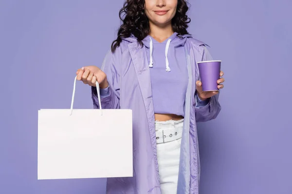 Vista recortada de mujer joven complacida en impermeable sosteniendo taza de papel y bolsa aislada en púrpura - foto de stock