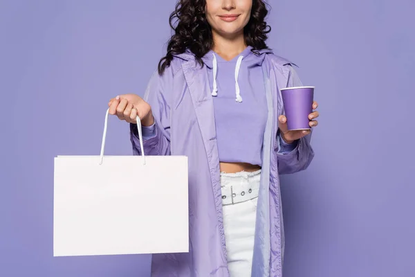 Vista recortada de mujer joven complacida en impermeable sosteniendo taza de papel y bolsa aislada en púrpura - foto de stock