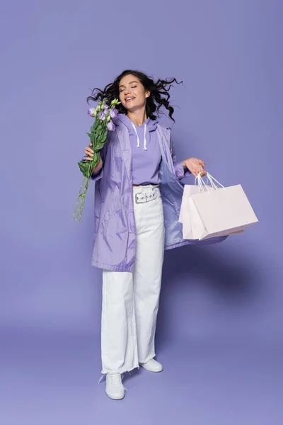 Heureuse jeune femme avec les yeux fermés tenant des sacs à provisions et des fleurs sur violet — Photo de stock