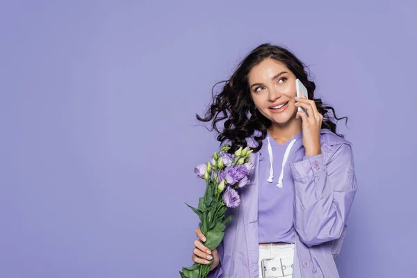 Glückliche junge Frau hält Blumen in der Hand und spricht auf dem Smartphone isoliert auf lila — Stockfoto