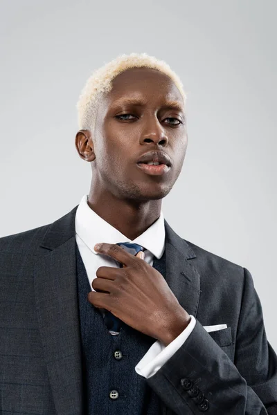 Portrait of blonde african american businessman in suit adjusting tie isolated on grey — Stock Photo