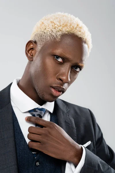 Portrait of blonde african american man in suit adjusting tie isolated on grey — Stock Photo