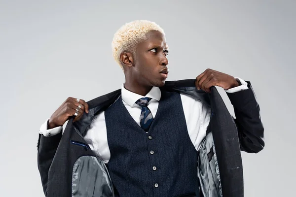 Blond afro-américain regardant loin et ajustant blazer isolé sur gris — Photo de stock