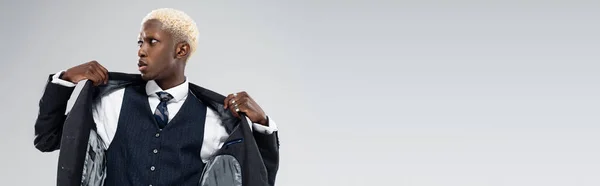 Biondo afroamericano che distoglie lo sguardo e regola il blazer isolato su grigio, banner — Foto stock