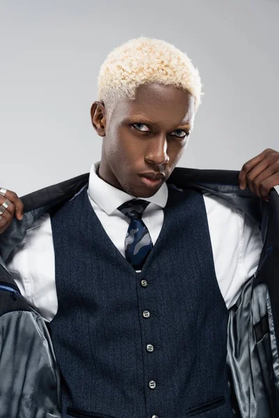 Blonde african american man looking at camera and adjusting blazer isolated on grey — Stock Photo