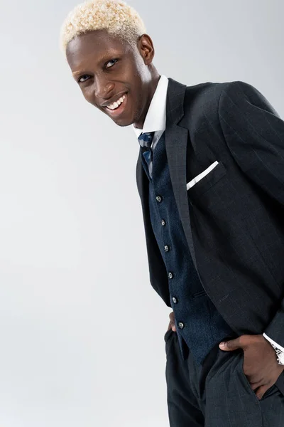 Alegre y joven hombre afroamericano en traje posando con las manos en bolsillos aislados en gris - foto de stock