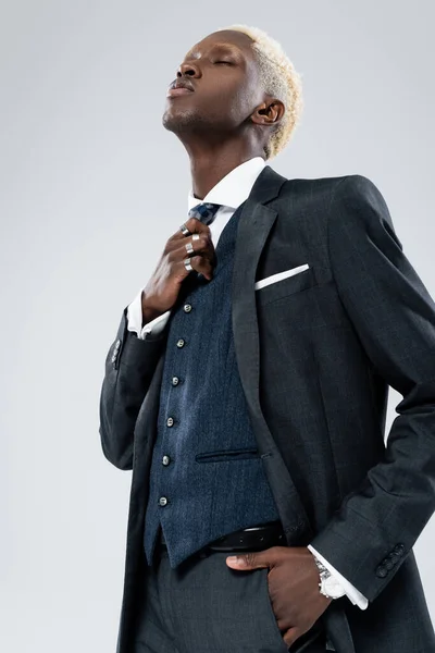Hombre afroamericano con estilo en ropa formal ajuste corbata y posando con la mano en el bolsillo aislado en gris - foto de stock