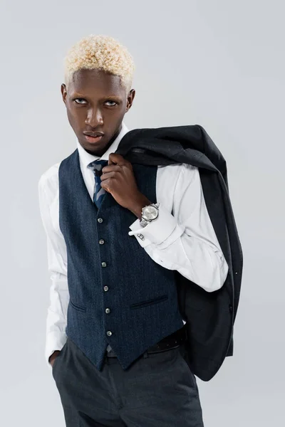 Hombre afroamericano rubio con ojos azules sosteniendo chaqueta y posando con la mano en el bolsillo aislado en gris - foto de stock