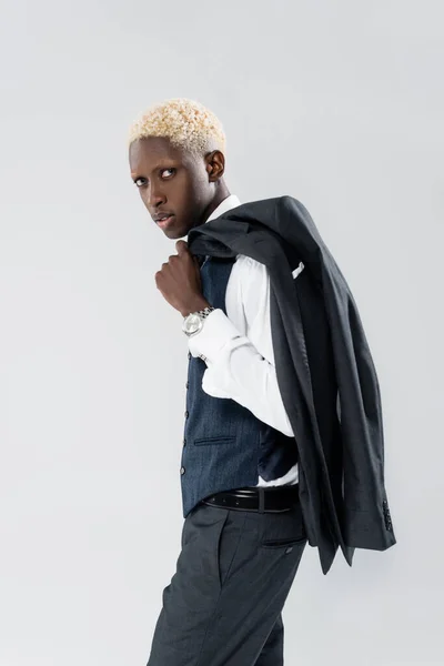 Biondo e giovane uomo afroamericano che tiene la giacca e distoglie lo sguardo isolato sul grigio — Foto stock