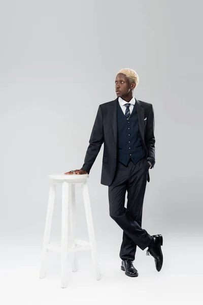 Full length of blonde african american man in suit standing with hand in pocket near white chair on grey — Stock Photo