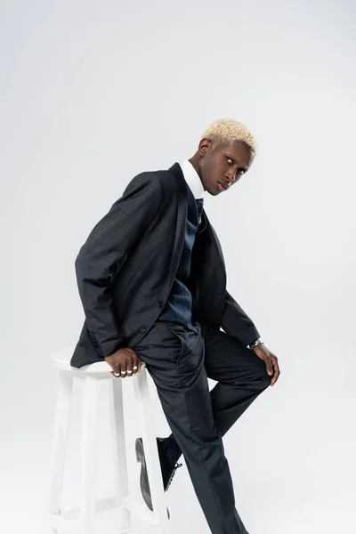 Young african american man in suit leaning on white chair isolated on grey — Stock Photo