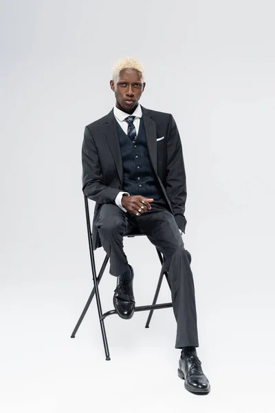 Full length of blonde african american man in suit sitting on chair on grey — Stock Photo
