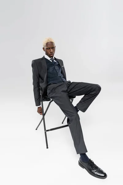 Full length of blonde and young african american man in formal wear sitting on chair on grey — Stock Photo