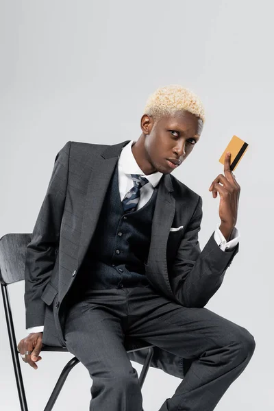 Loira e jovem empresário afro-americano de terno sentado na cadeira e segurando cartão de crédito isolado em cinza — Fotografia de Stock
