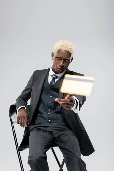 Blonde african american businessman in suit sitting on chair and throwing blurred credit card isolated on grey — Stock Photo