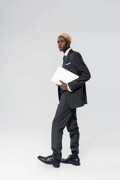 Full length of blonde african american businessman in suit holding laptop on grey — Stock Photo