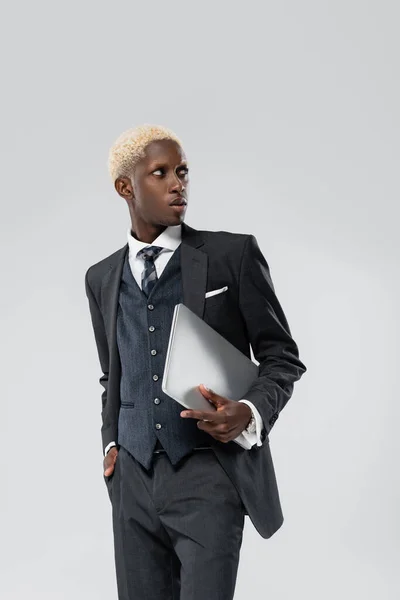 Homme d'affaires afro-américain blonde en costume tenant un ordinateur portable et posant la main dans la poche isolé sur gris — Photo de stock