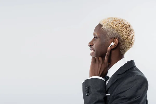 Side view of happy african american man adjusting wireless earphone isolated on grey — Stock Photo